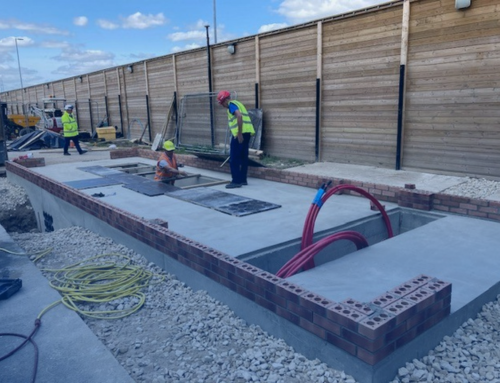 Construction workers are seen on a building site, working on a concrete foundation surrounded by brickwork. Tools and materials are scattered around, indicating ongoing construction activity.