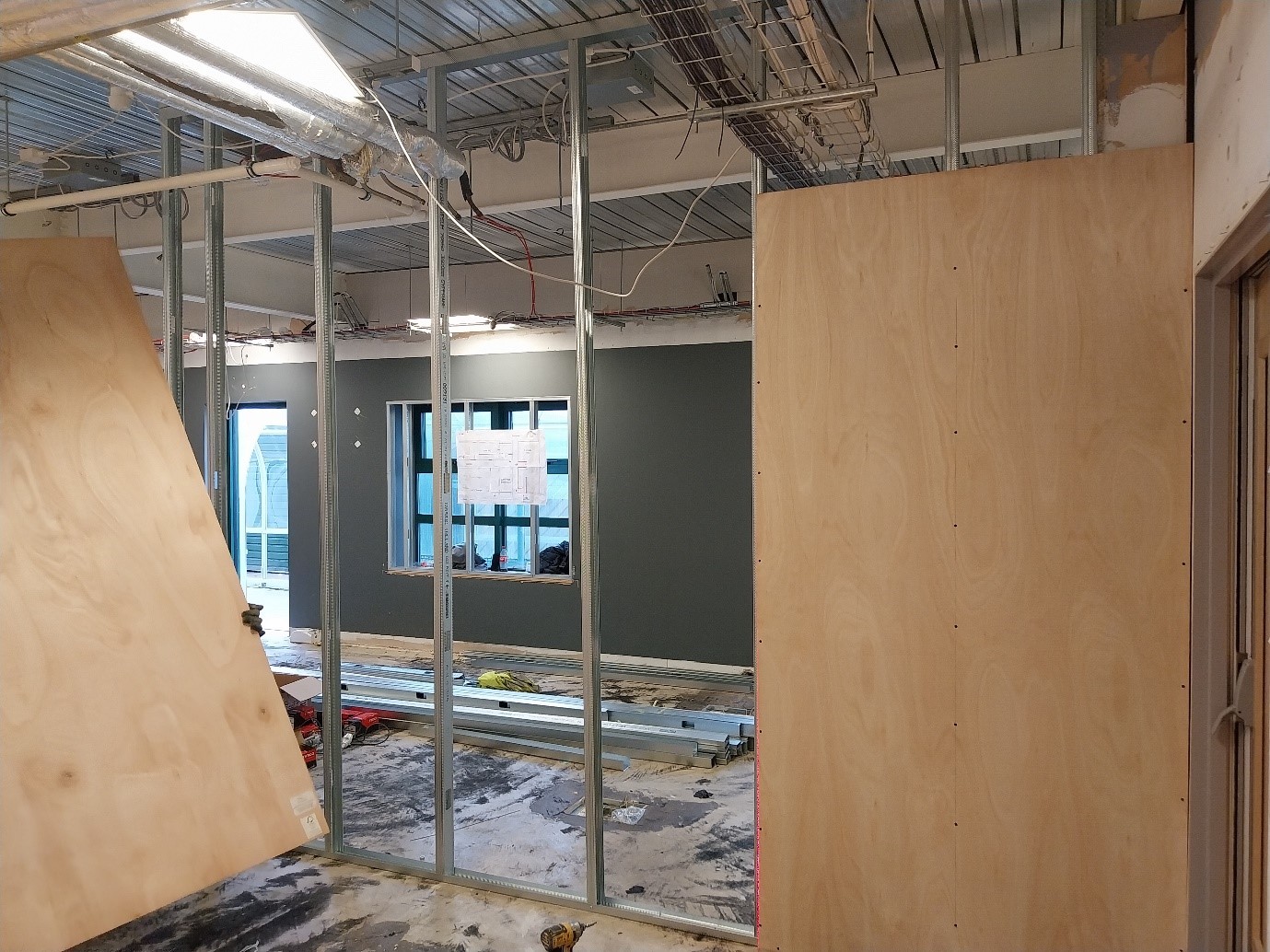 The image depicts an interior construction site with metal framing and plywood panels. A dark green wall is partially visible, along with tools and materials scattered on the floor.