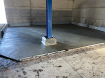 A freshly poured concrete floor is visible, with a blue support column in the center. The surrounding area shows unfinished surfaces and construction materials.
