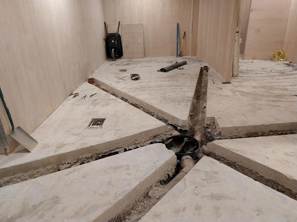 Picture12 A construction site features a concrete floor with several trenches and a central drainage system. Tools and equipment are scattered around, indicating ongoing work in the space.