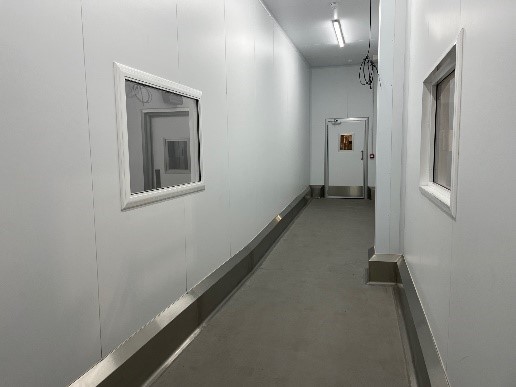 A narrow, well-lit corridor features smooth, white walls and a concrete floor. There are two windows on the left side and a door at the far end, suggesting a clean and controlled environment.