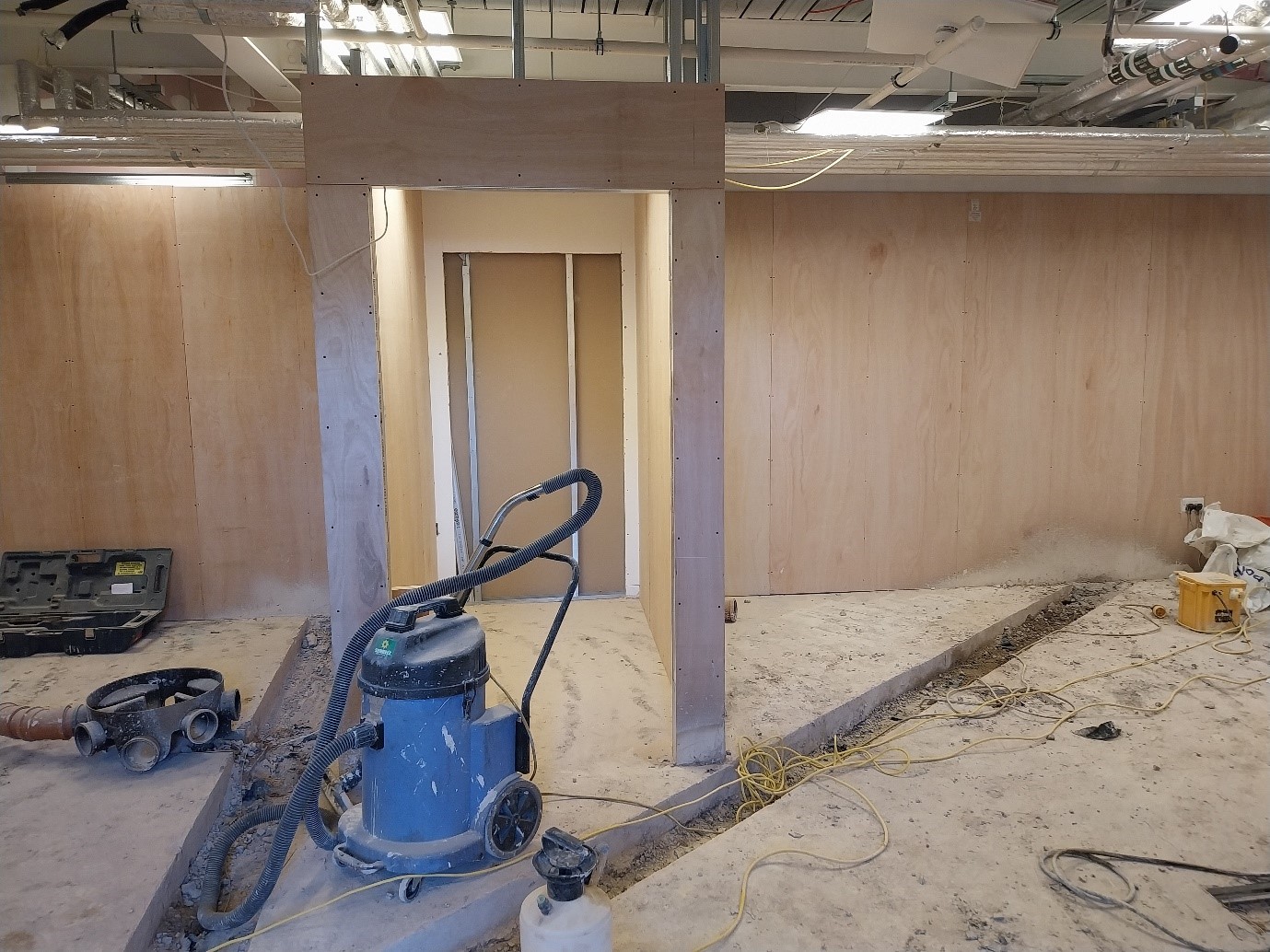 A construction site is depicted, featuring wooden walls and an unfinished doorway. Various tools and equipment, including a vacuum cleaner and electrical cords, are scattered across the dusty floor.