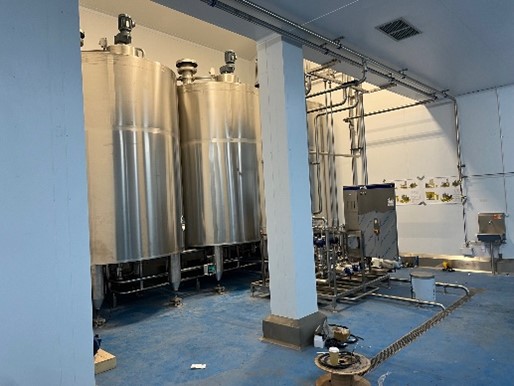 The image features a clean industrial space with large stainless steel tanks and various pipes and machinery. The floor is blue, and there are some tools and equipment scattered around, indicating an active work environment.