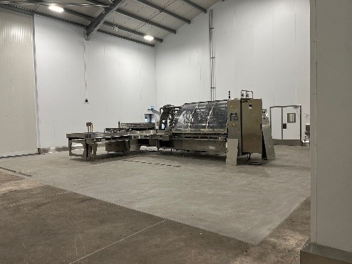 A spacious industrial room features a large, modern machine surrounded by concrete flooring. The walls are white, and the area is well-lit, indicating a clean and organized workspace.