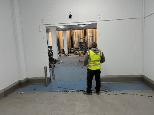 A person wearing a high-visibility vest stands in front of an open doorway, looking into a large, industrial space with metal tanks in the background. The floor is blue and appears to be in a construction or installation phase.