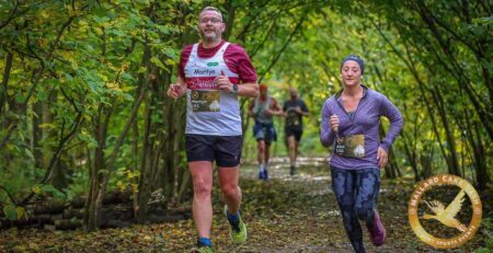 Two runners are seen participating in a race along a tree-lined path, surrounded by autumn foliage. One runner is wearing a red and white shirt, while the other is in a purple top, both displaying their race numbers.