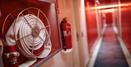 A fire hose is mounted on a wall in a brightly lit corridor, with a fire extinguisher visible nearby. The hallway features red walls and soft lighting, creating a striking visual contrast.