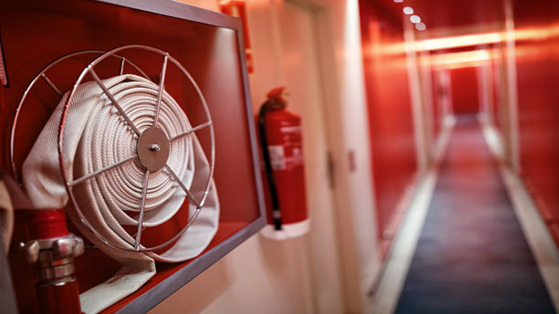 A fire hose is mounted on a wall in a brightly lit corridor, with a fire extinguisher visible nearby. The hallway features red walls and soft lighting, creating a striking visual contrast.