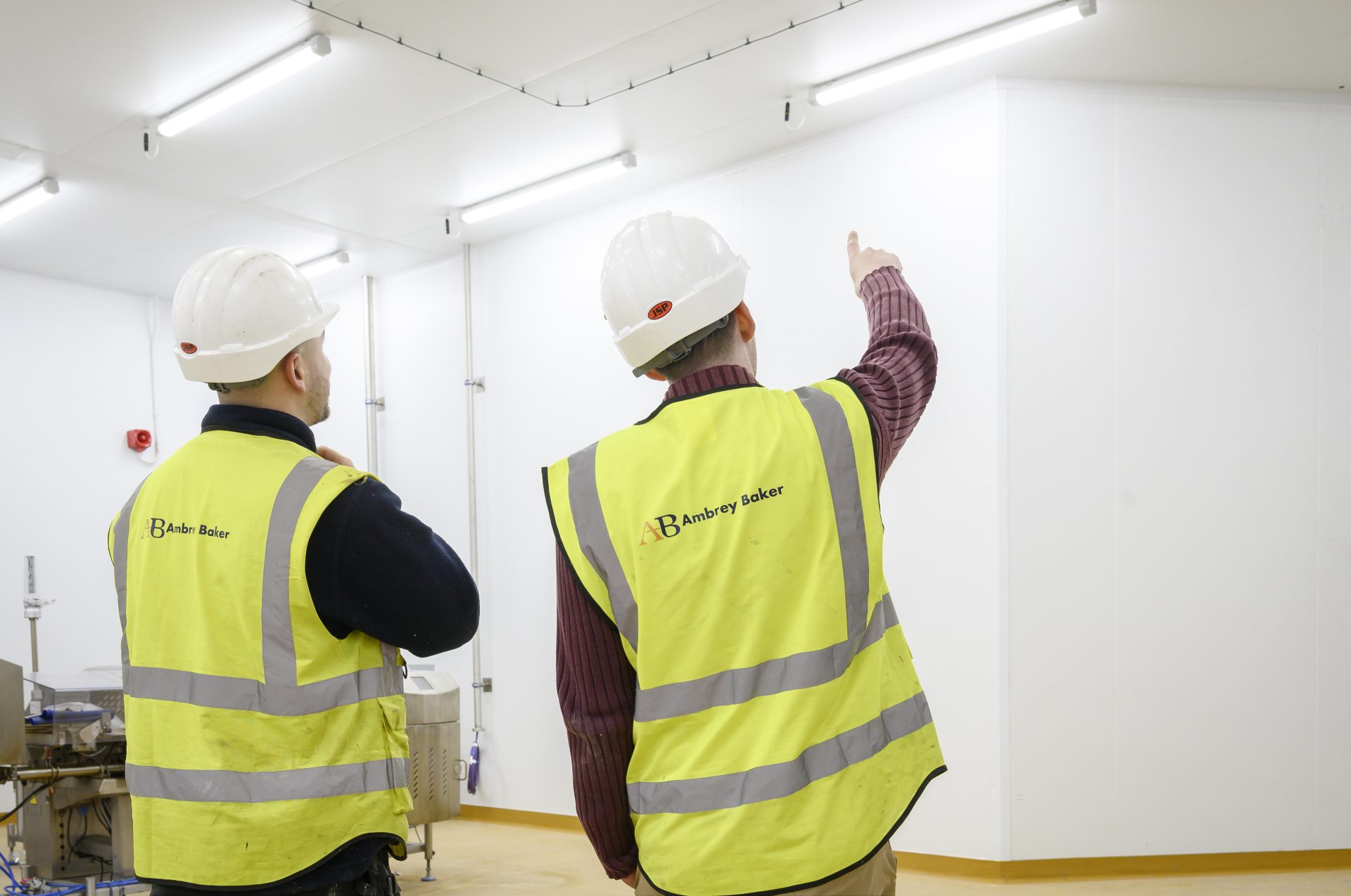 peteralvey.com-2024-04-05-76 Two individuals in safety vests and hard hats are discussing something in a well-lit, industrial space. One person is pointing towards the ceiling, indicating an area of interest.